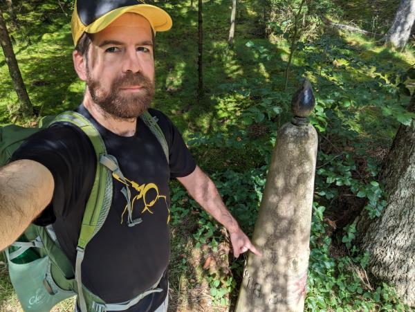 A man pointing at a waist-height concrete post in the middle of a forest.