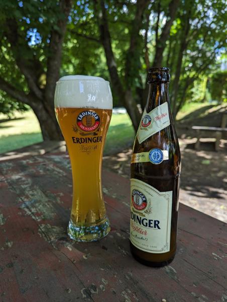 A photo of a glass full of Erdinger beer with the empty bottle sitting on a picnic table.