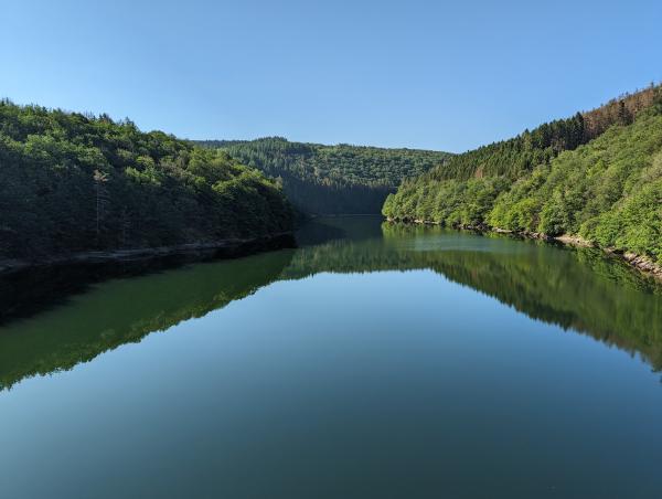 A view across a wide lake from high up with forested hills in the background.