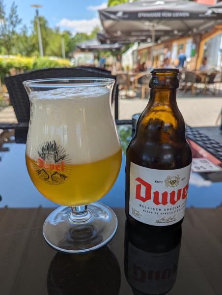 A photo of a tulip shaped glass full of Duvel beer and the empty bottle sitting on a restaurant table.