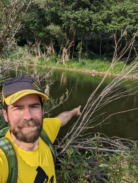 A man standing in a thicket pointing to a river in the background.