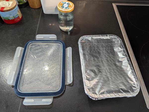 A photo of a dark countertop with the foil covered baking dish, a clear plastic lid and a small glass jar of water sitting on it.