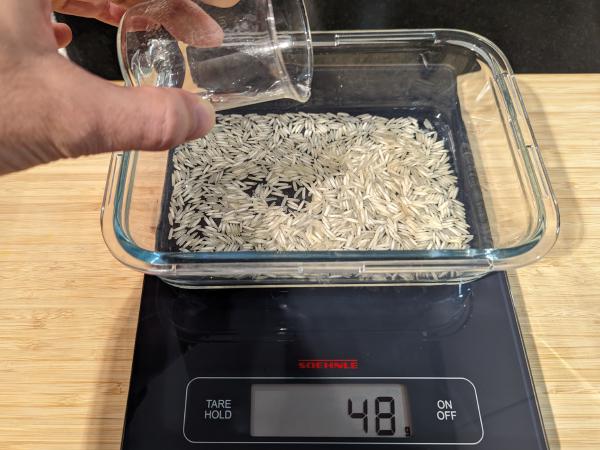 A photo of the same baking dish now with water in the rice and the scales reading 48 g. There is a hand holding a small glass beaker with water in it hovering over the dish.