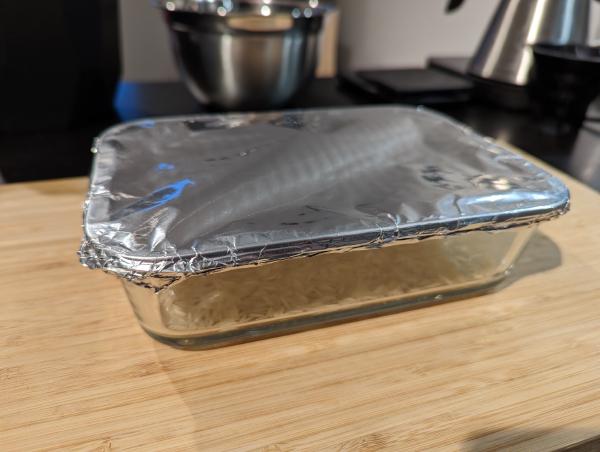 Another photo of the baking dish with rice and water in it sitting on a wooden cutting board. The baking dish has been covered in aluminium foil.