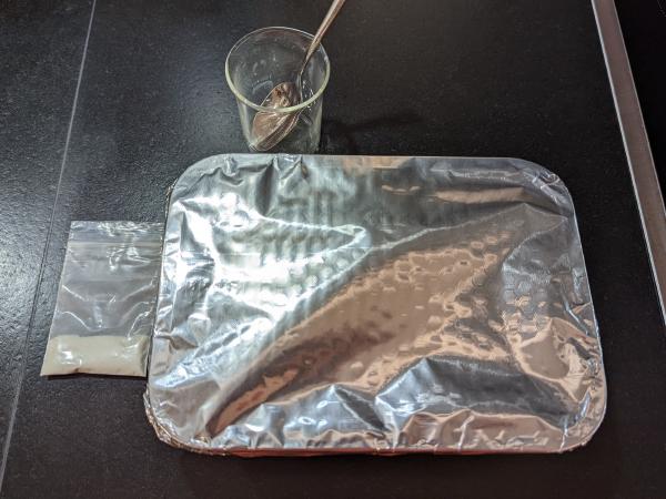 A photo of a dark countertop with a number of items sitting on it. A glass beaker with a teaspoon in it, the covered baking dish and a small plastic bag of beige powder.