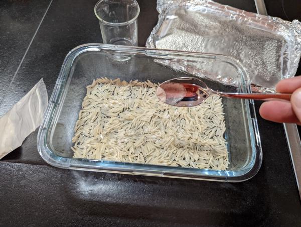 The same setup, but the foil has been removed from the baking dish, the bag of beige powder opened and the teaspoon has been used to scatter the powder over the surface of the rice.
