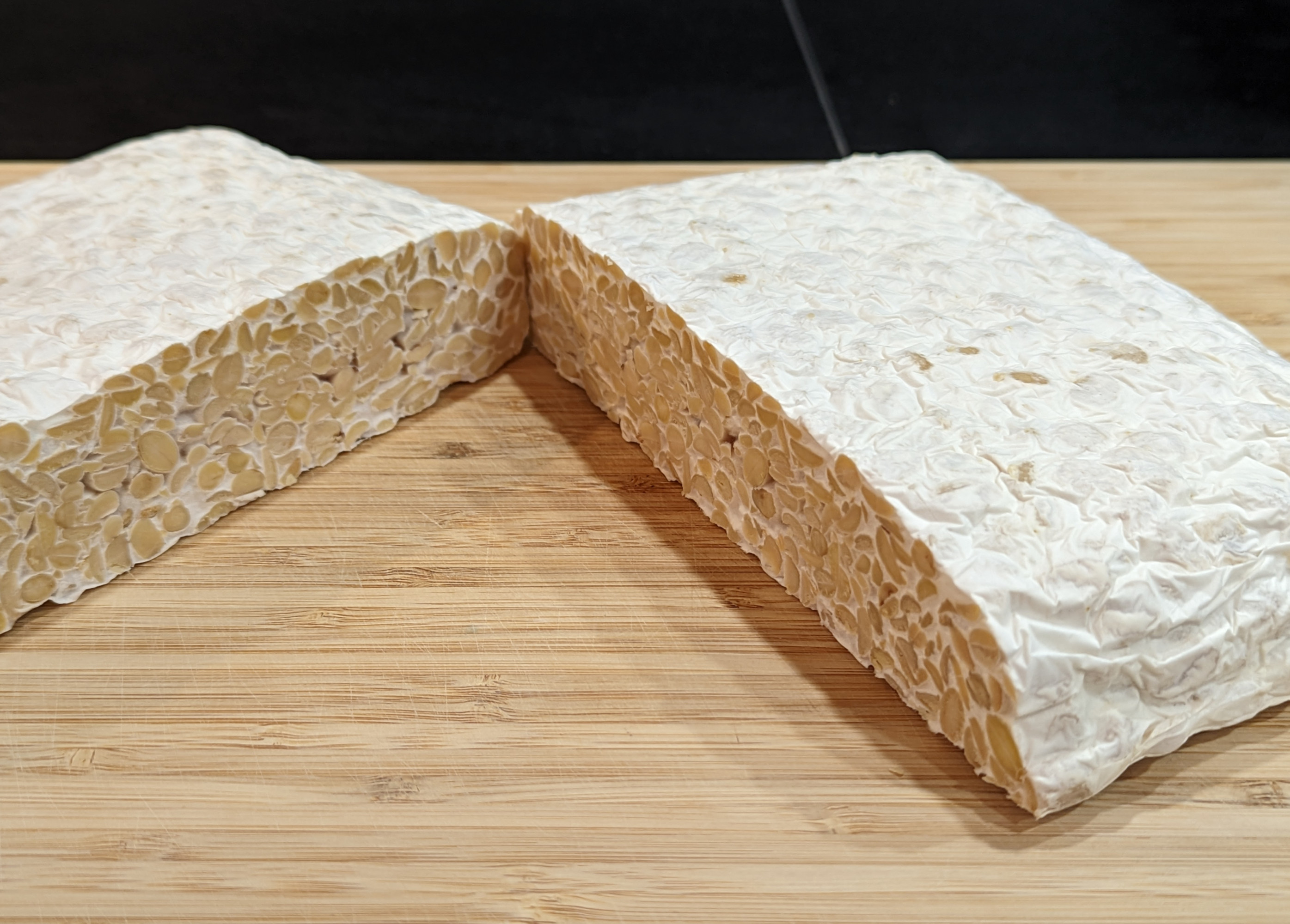 A photo of a white mould-covered cake of soybeans sliced in two, sitting on a wooden cutting board.